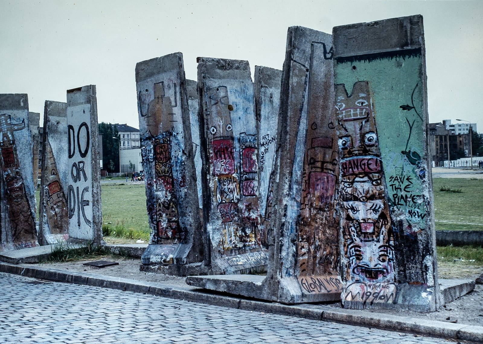 Colour photo of a row of graffiti-covered Wall segments left standing on a cobbled pavement beside a grassy open space, with apartment buildings in the distance.