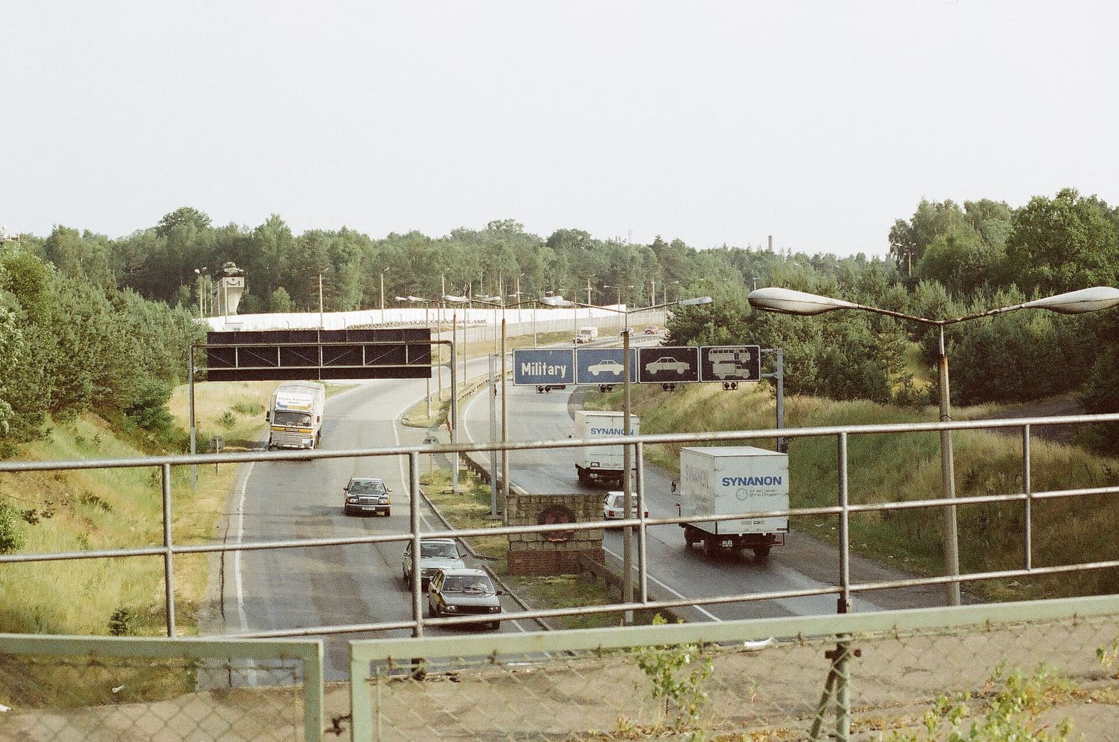 Colour photo from a bridge railing over a divided autobahn with lorries and cars, overhead lane signs including 'Military', and a white wall and watchtower in the distance.