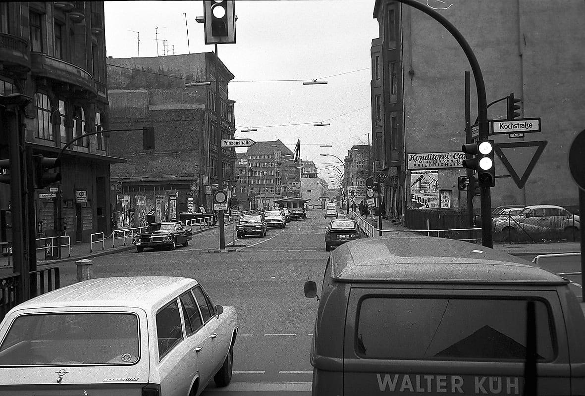 Black-and-white street scene with cars and traffic lights at a junction, the Allied checkpoint hut in the middle of the road and the border installations beyond.