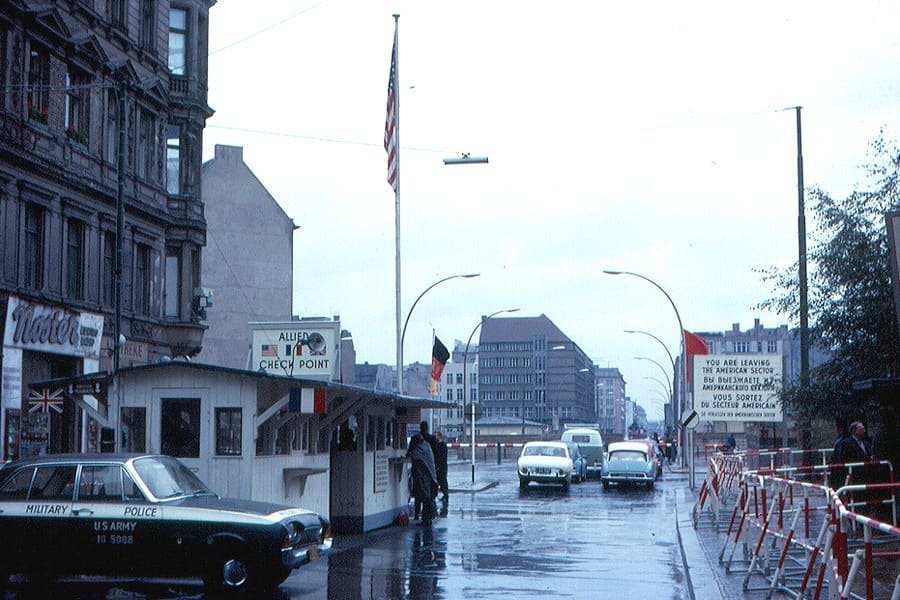 Colour photo of the Allied checkpoint hut in the middle of Friedrichstraße, a US Army military police car beside it and a road leading east.