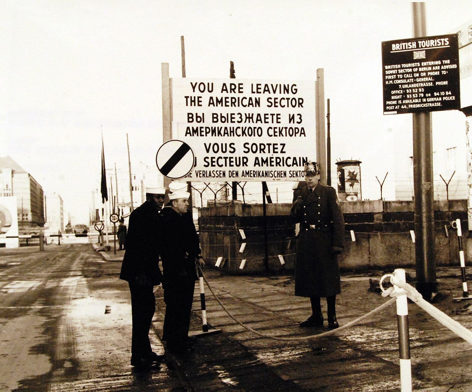 Black-and-white photo of uniformed men beside a large four-language sign reading 'You are leaving the American sector' in a snowy street.
