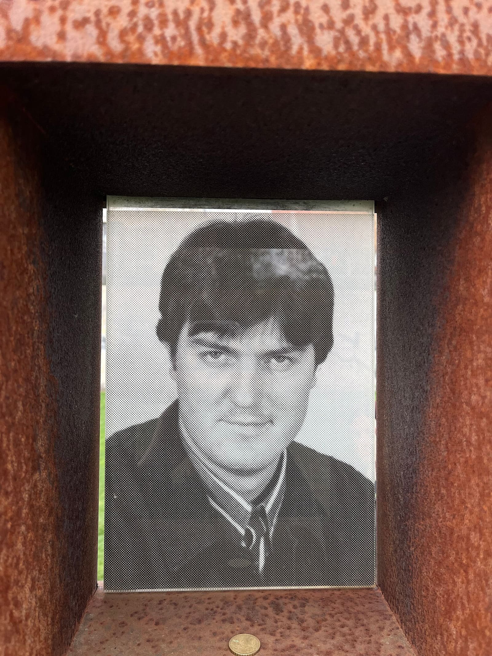 A portrait window set into a rust-coloured steel memorial wall, showing a photograph of a young man