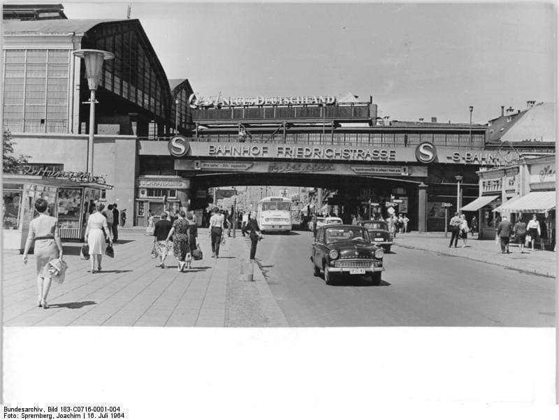 Black-and-white street scene with pedestrians and cars in front of a railway bridge signed 'Bahnhof Friedrichstrasse' and a large glass train hall.