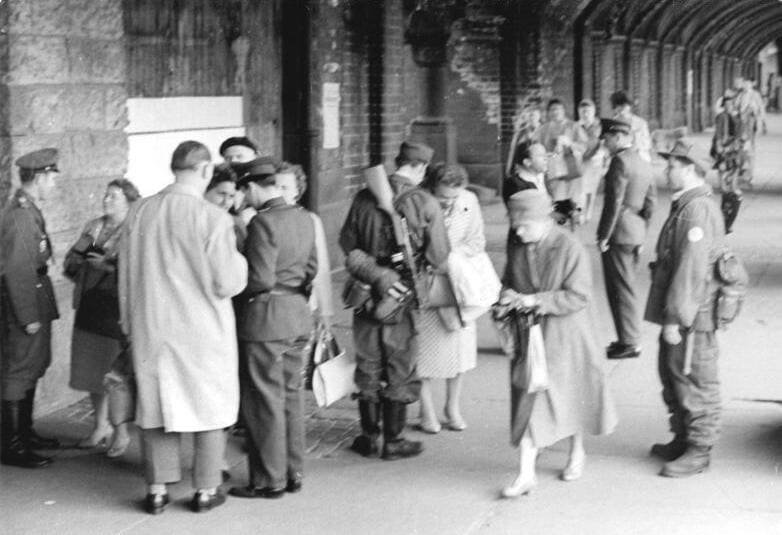 Black-and-white photo under the brick arcade of a bridge: uniformed East German police and an armed man in combat dress check the papers of men and women carrying handbags and coats.