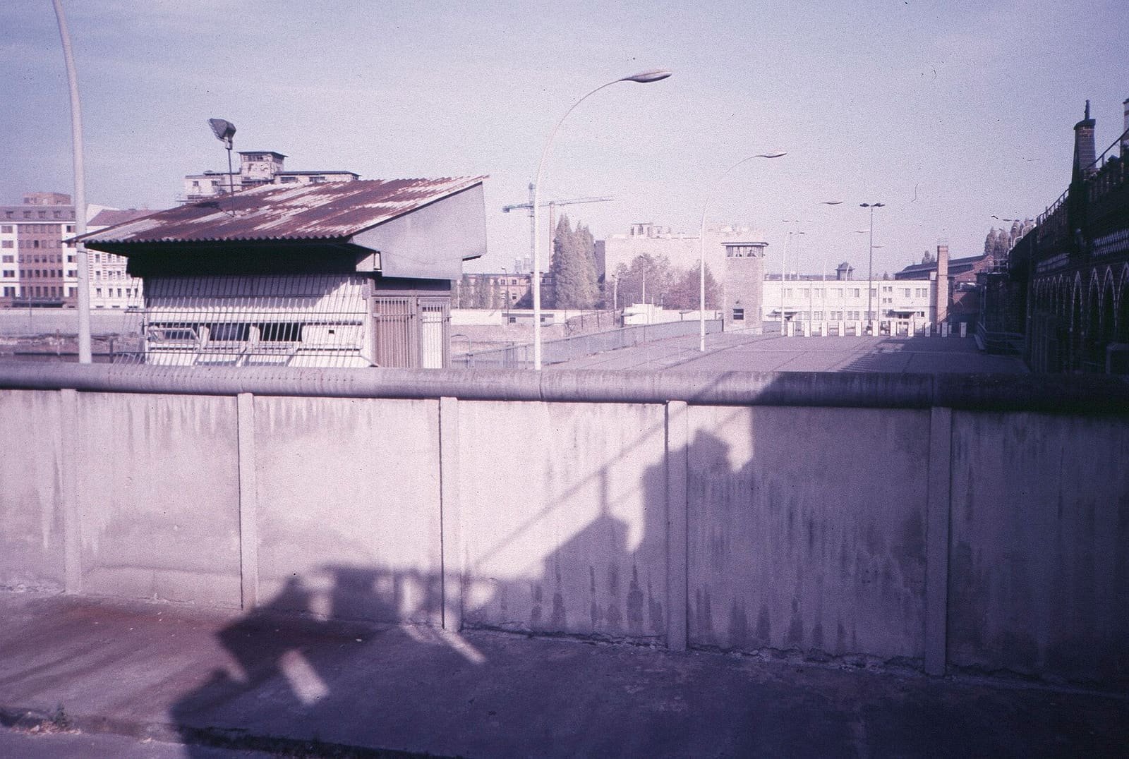 Colour photo over a concrete wall: an empty bridge roadway with lamp posts leads to a checkpoint hut and a watchtower, with the brick arcades of the bridge on the right and buildings beyond.