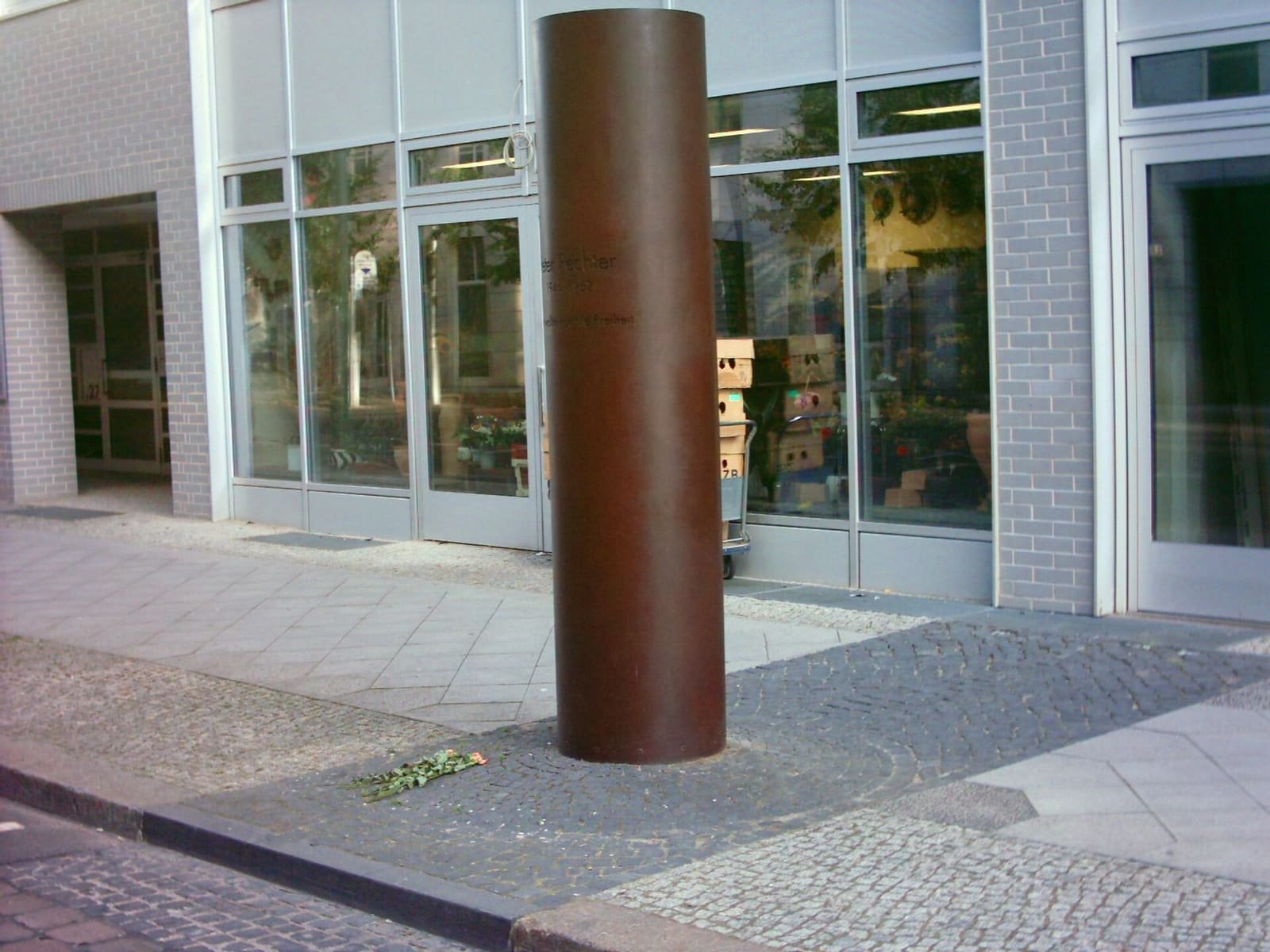 A slender dark memorial column on the pavement of a city street lined with modern buildings