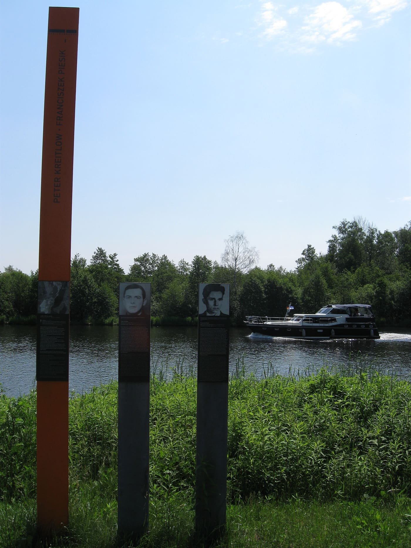 An orange stele and two grey posts with portrait panels on a grassy riverbank, with the Havel and a passing motorboat behind.