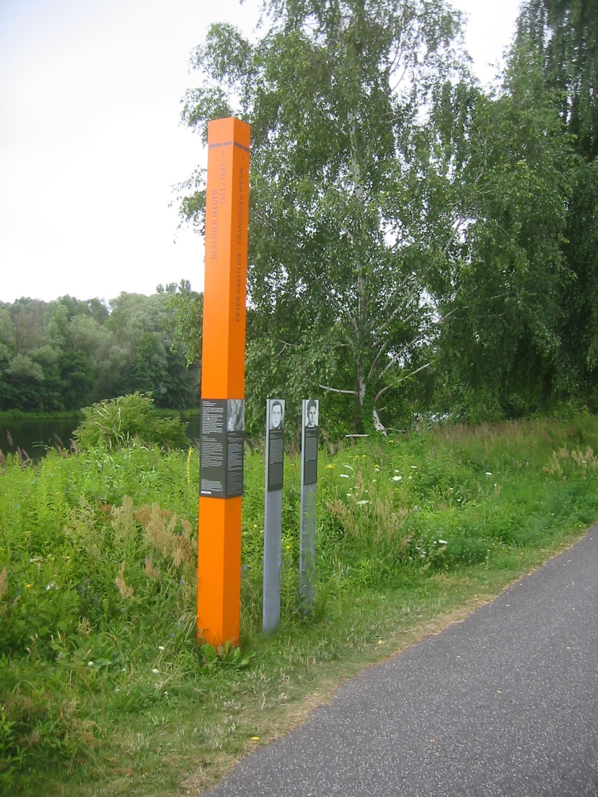 An orange stele and two slim grey posts standing in long grass beside a paved cycle path, with trees and water behind.