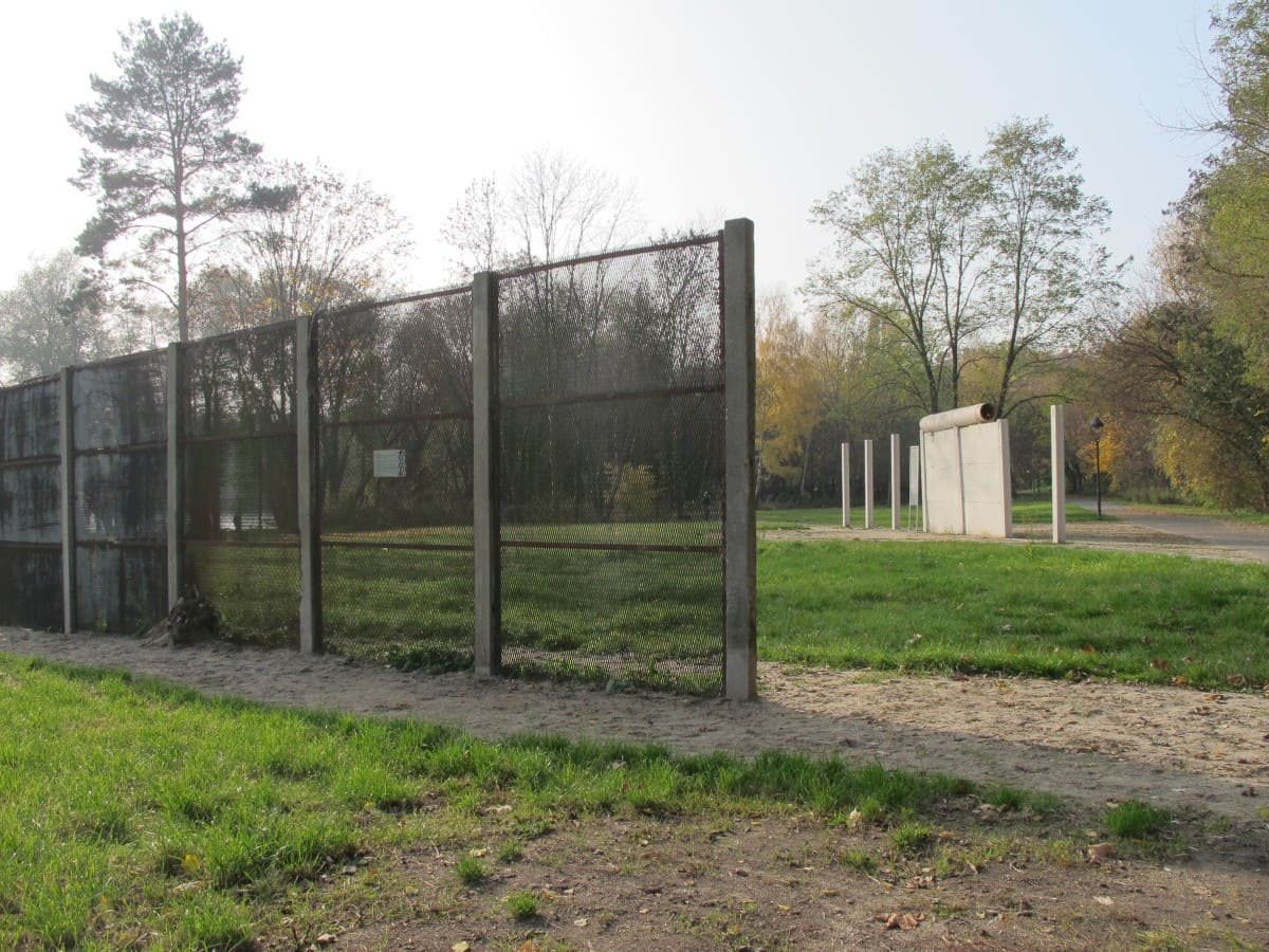 A row of concrete posts holding rusty mesh fence panels on a sandy path, with two Wall segments and white posts standing on a lawn behind.