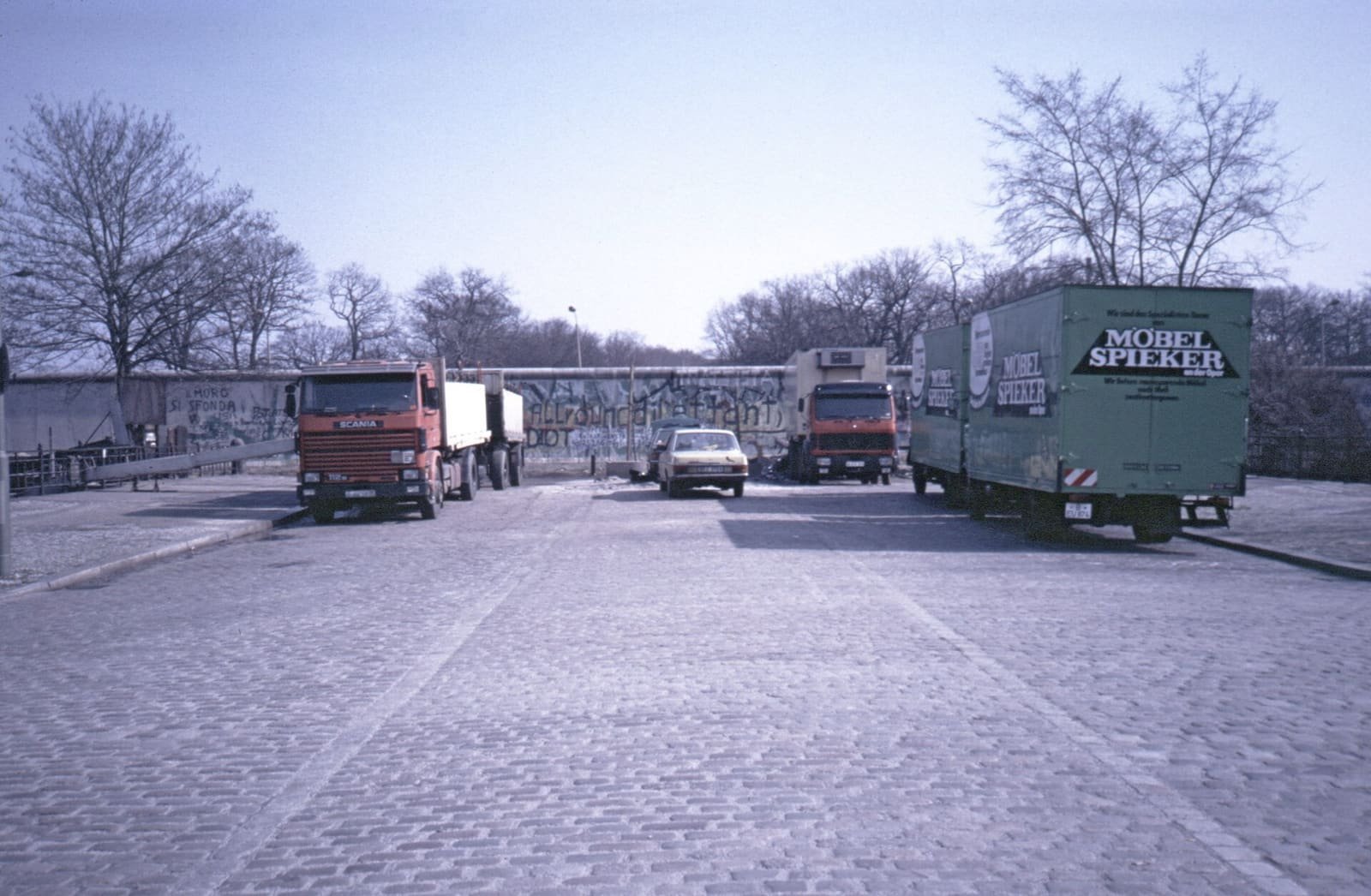 A cobbled square with parked lorries and a car ends at a graffiti-covered concrete wall stretching across the road, with bare trees behind it.
