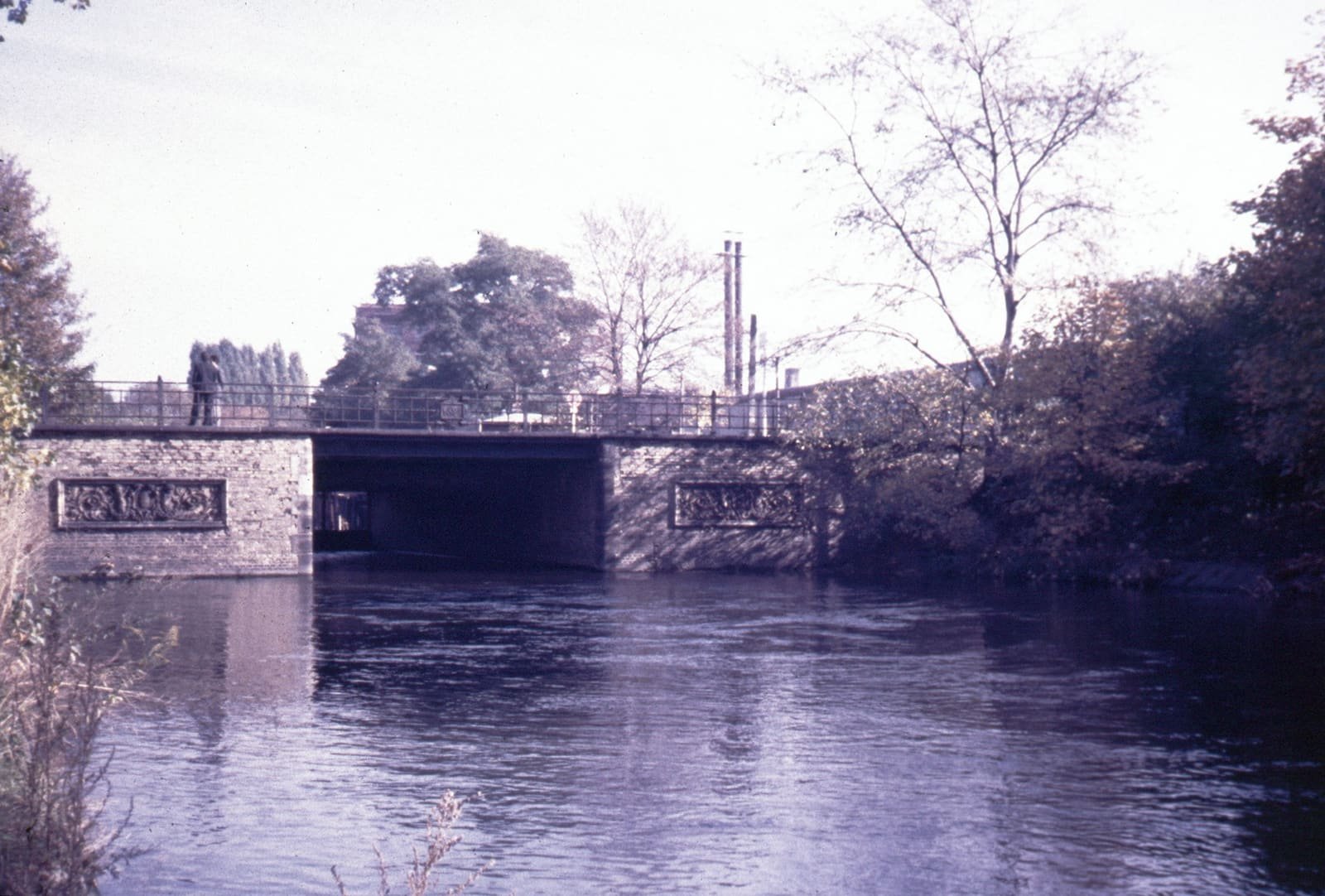 A low brick bridge with decorative panels spans a calm canal lined with autumn trees; a person stands at the railing.