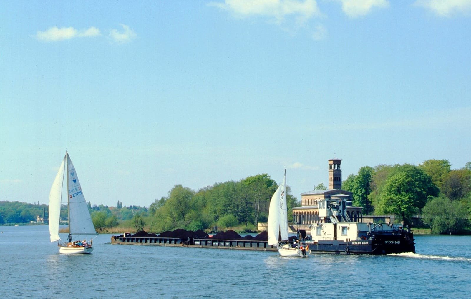Colour photo: sailing boats and a GDR push-tug with a barge on a wide river; on the far wooded bank stands a church with a tall campanile.