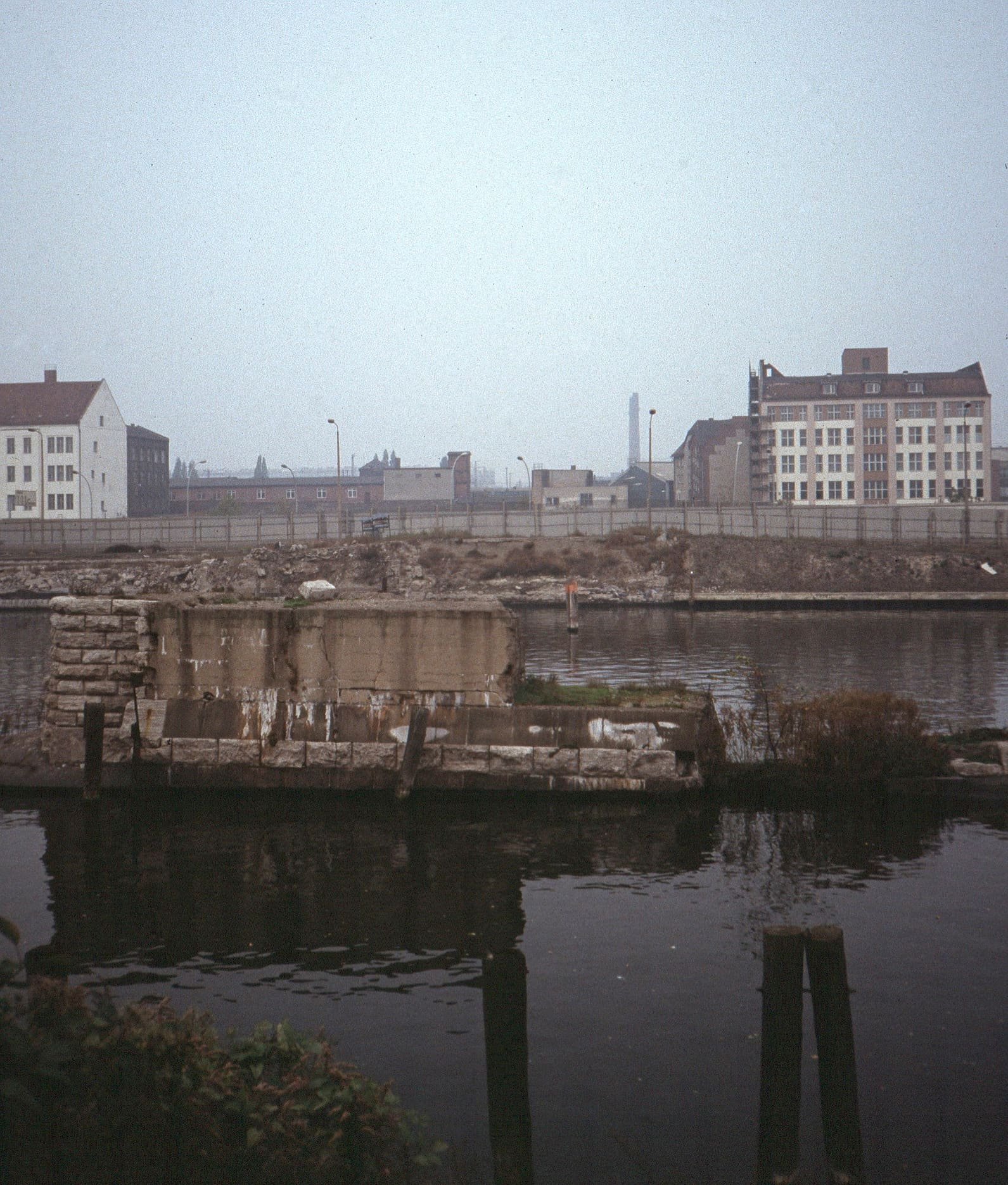A stone bridge pier in dark water, with a wall and industrial buildings on the far riverbank under a grey sky.