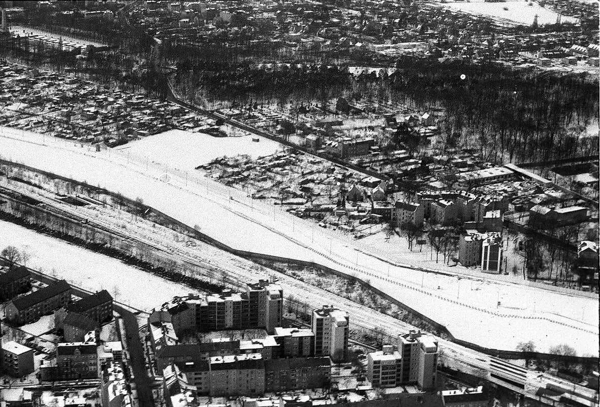 Black-and-white aerial photo of a wide, snow-covered border strip running diagonally between allotments and houses above and apartment blocks and a railway below.