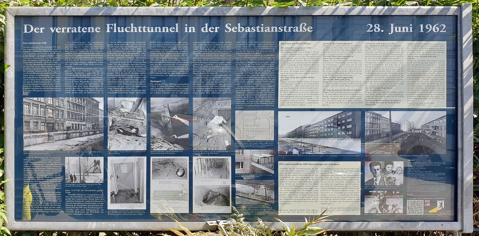 Wide blue outdoor information board with German text columns and many black-and-white photos of the tunnel and people involved, set among plants.