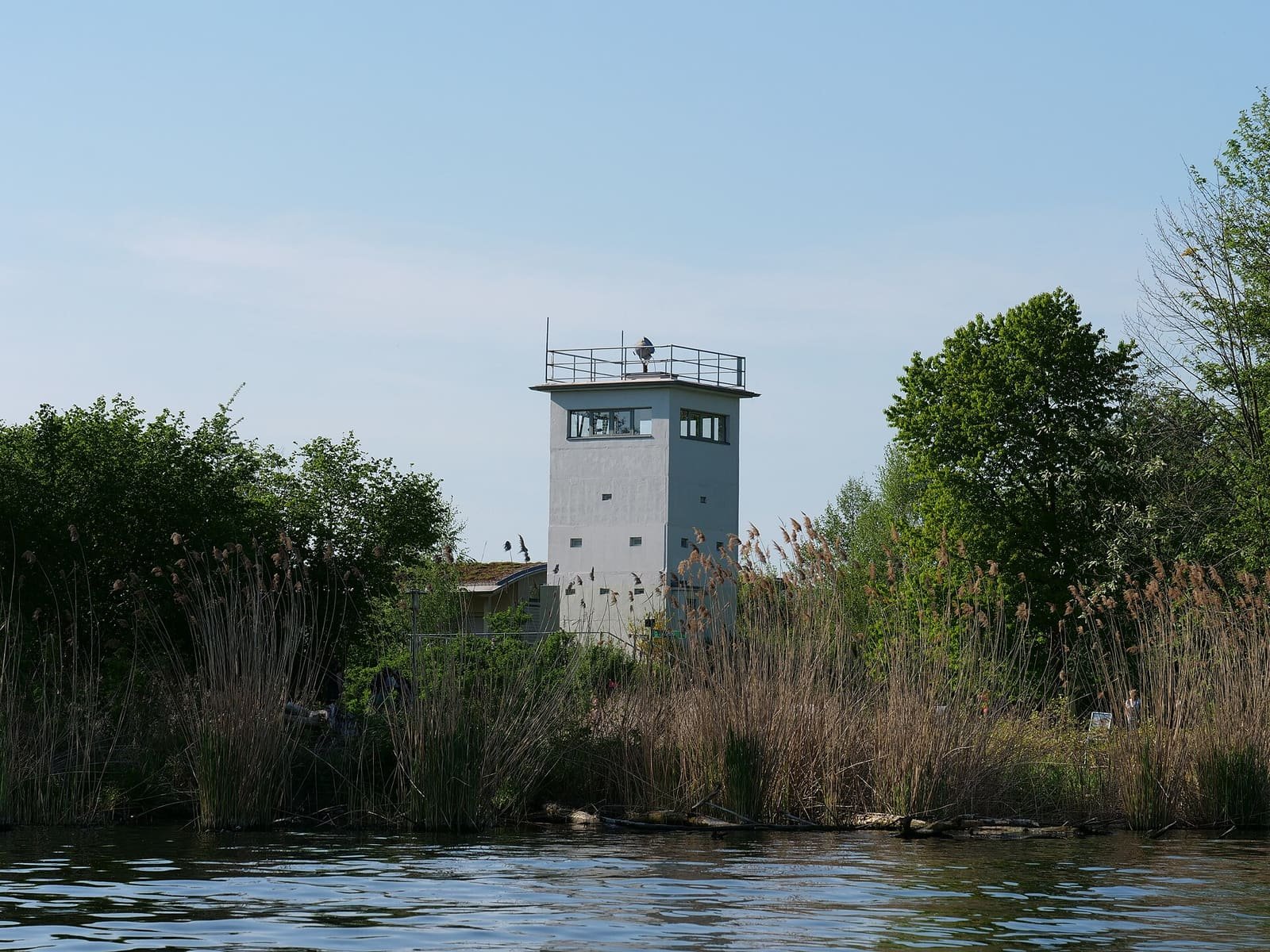 A square grey concrete watchtower with a railed roof platform rises behind tall reeds and trees on a riverbank, under a clear blue sky.