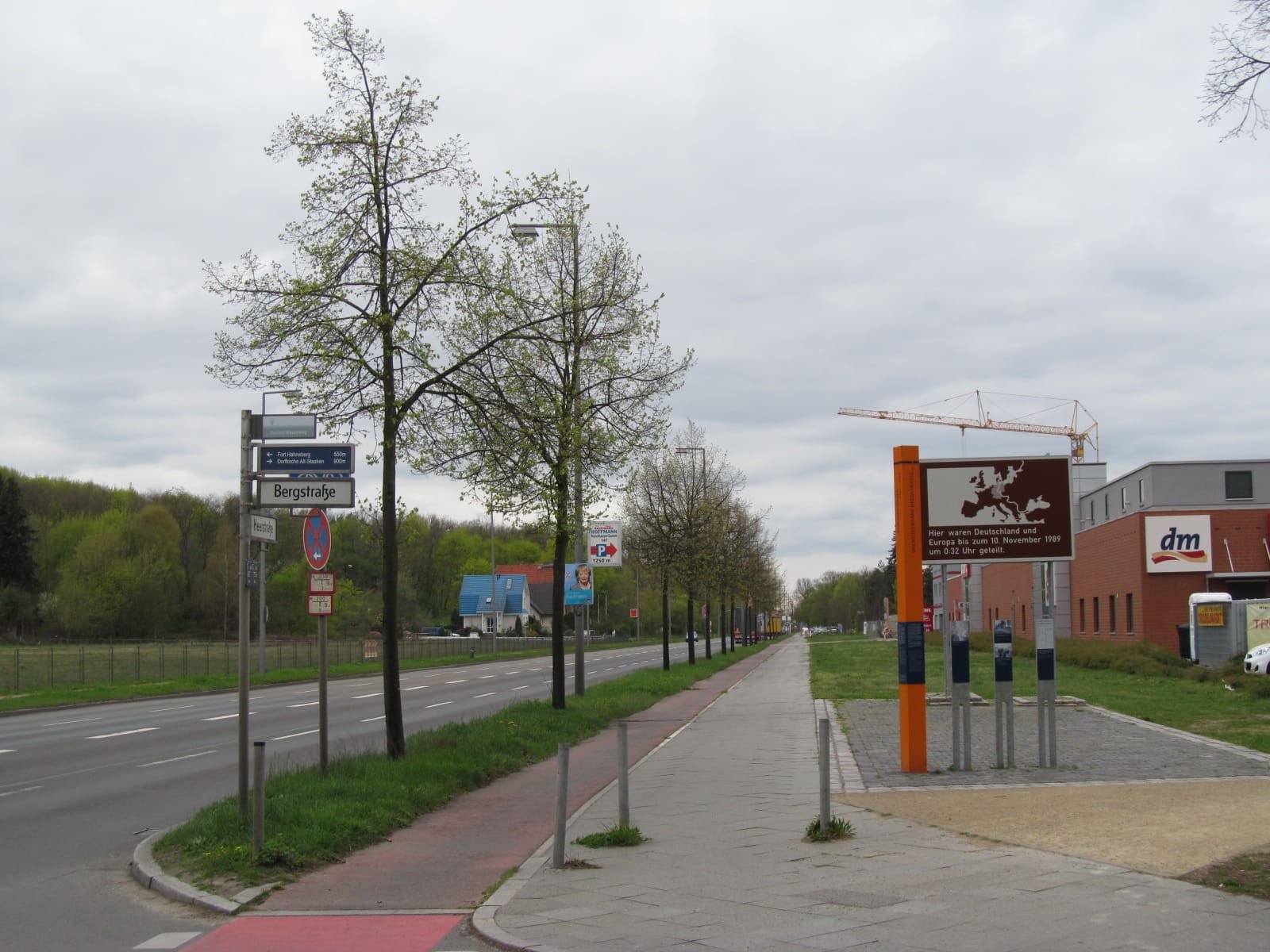 A wide road with a tree-lined cycle path; at right, an orange post, information steles and a brown sign with a map of Europe stand on a paved area.