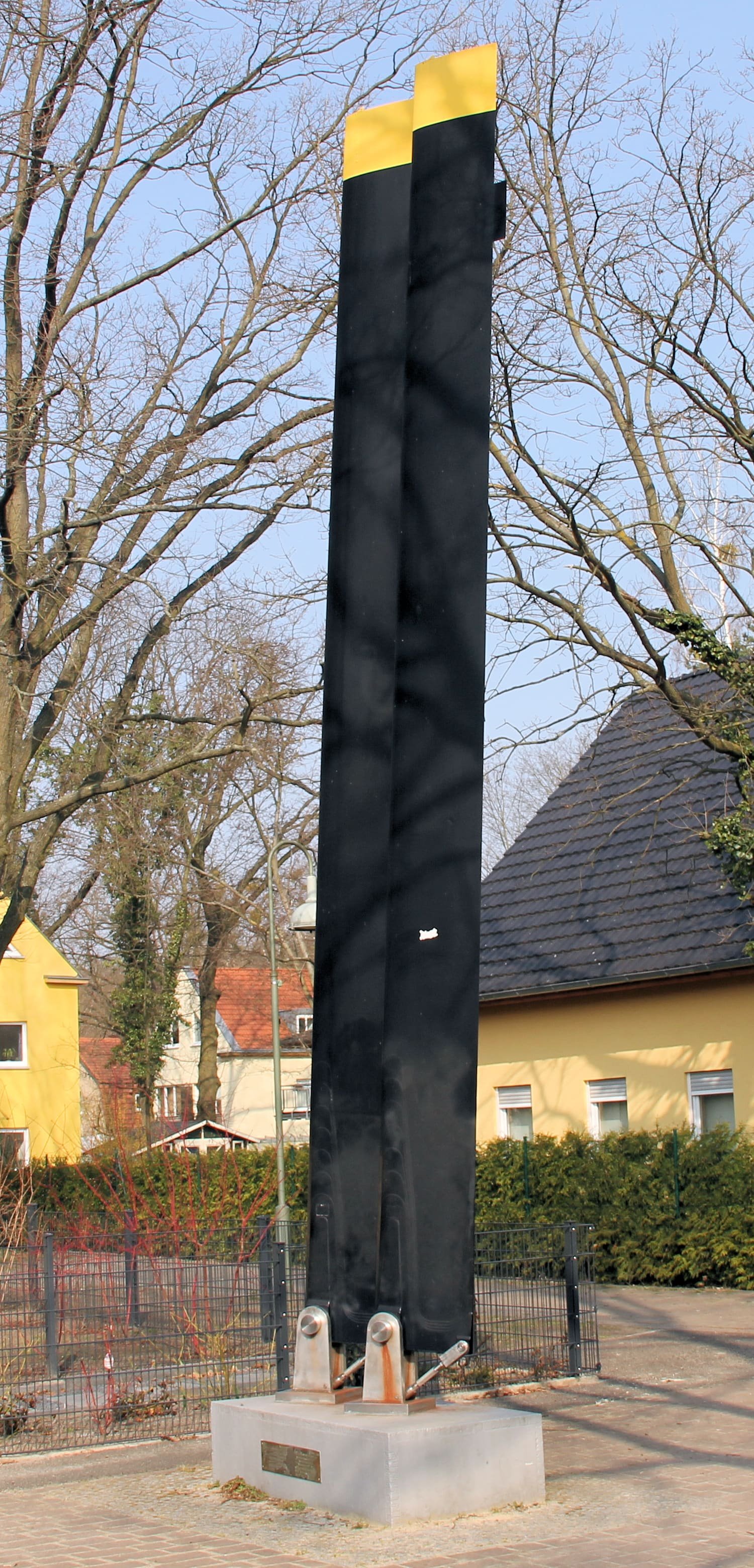 Two tall black helicopter rotor blades with yellow tips stand upright on a concrete plinth in front of houses and bare trees.