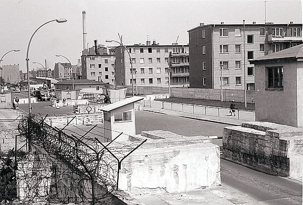 Black-and-white photo looking over barbed wire and a concrete wall to a checkpoint hut and road, with new blocks of flats on the East Berlin side.