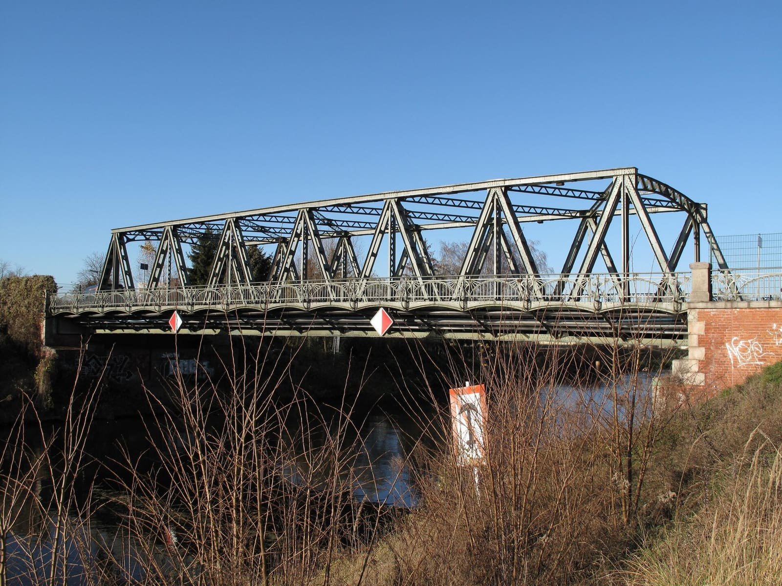 Colour photo of a steel truss road bridge over a canal under a clear blue sky, with dry reeds in the foreground and a brick abutment on the right.