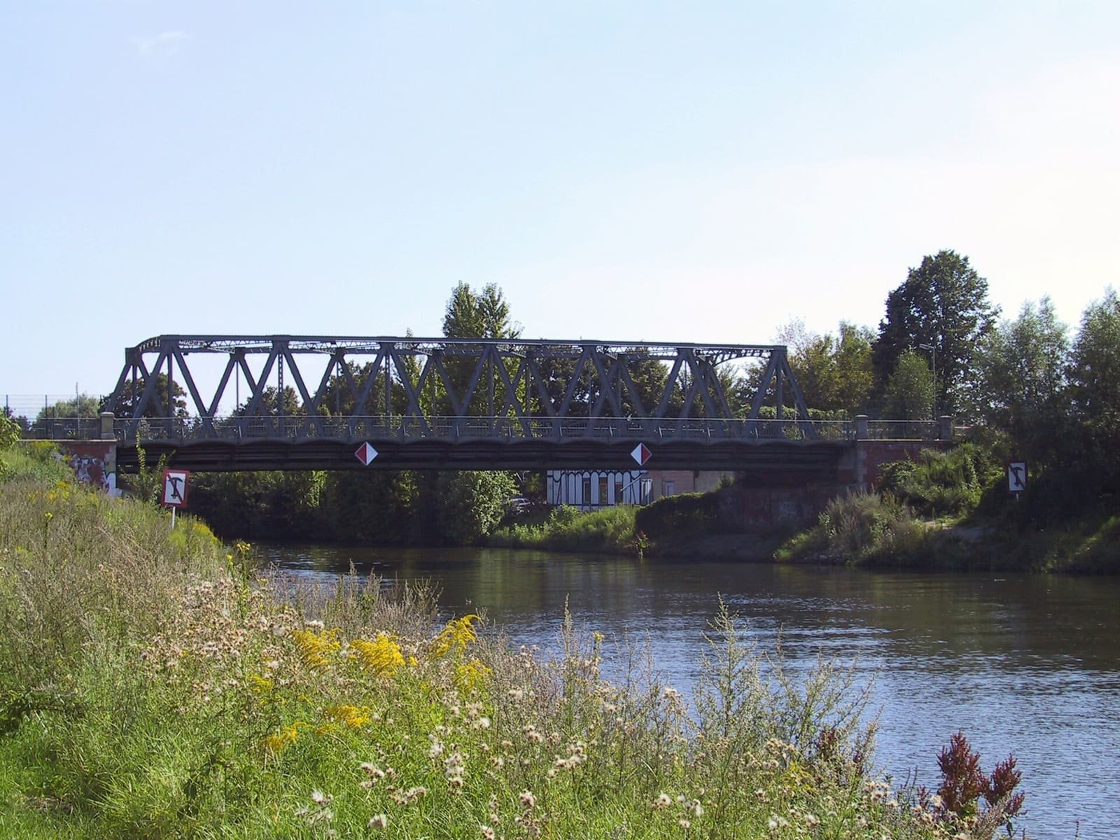 Colour photo of a steel truss bridge spanning a calm canal, with wildflowers on the near bank and trees on the far side.