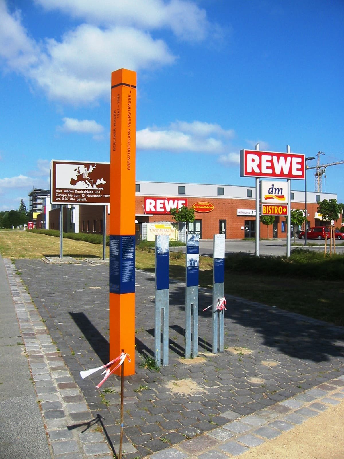 A tall orange stele and three smaller information panels on a paved area, with a supermarket behind.
