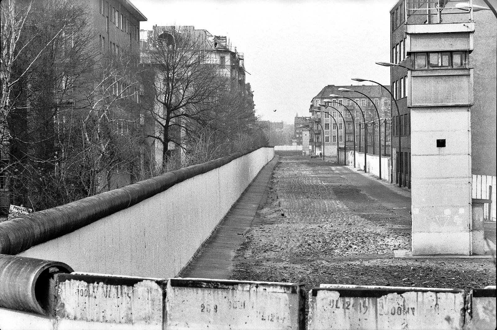 Black-and-white photo from the top of the Wall: a long concrete wall runs beside a sandy patrol strip with lamp posts and a watchtower on the right, between apartment buildings.