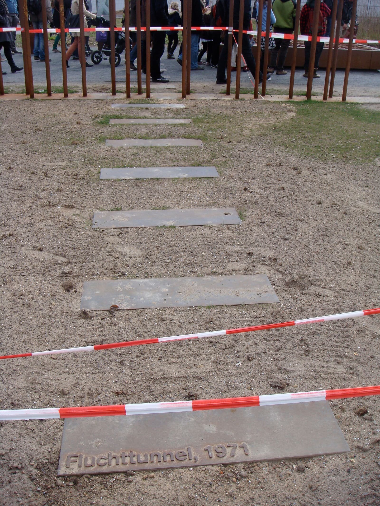 A row of steel plates in sandy ground leading to a row of rusted steel rods, the nearest plate inscribed 'Fluchttunnel, 1971'; visitors stand behind the rods.