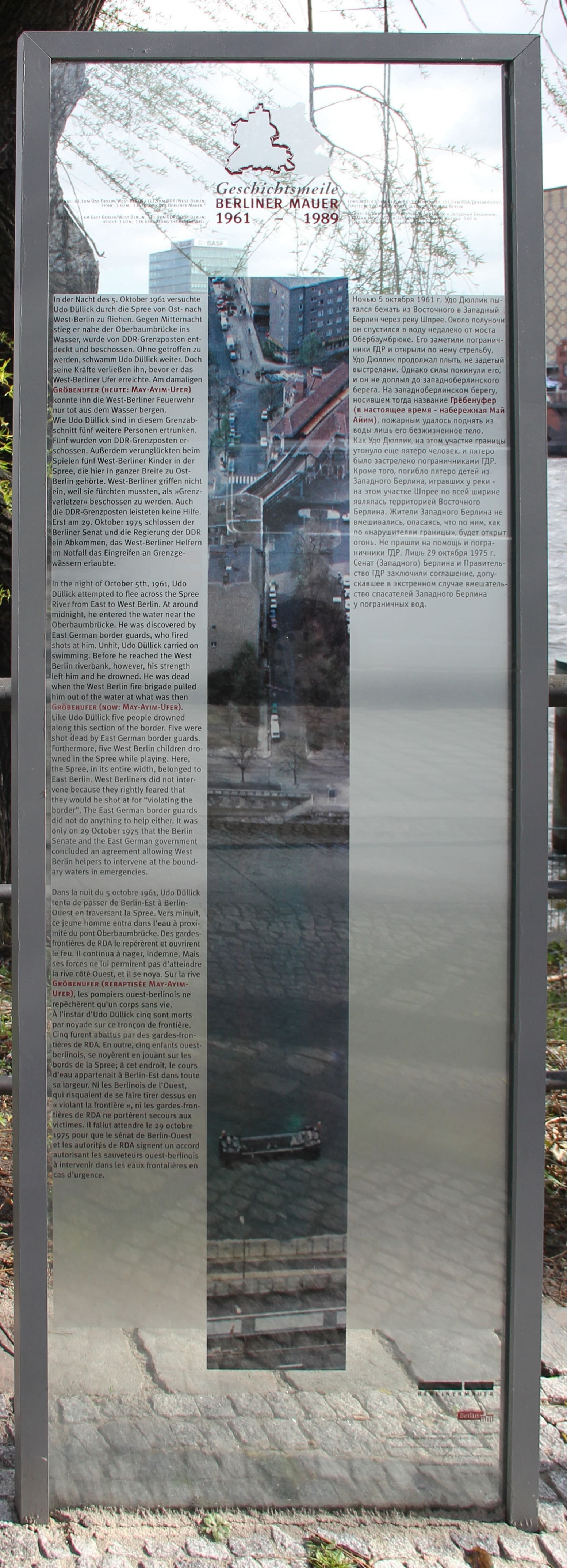 A tall memorial plaque with text commemorating Udo Düllick, standing by the river bank.