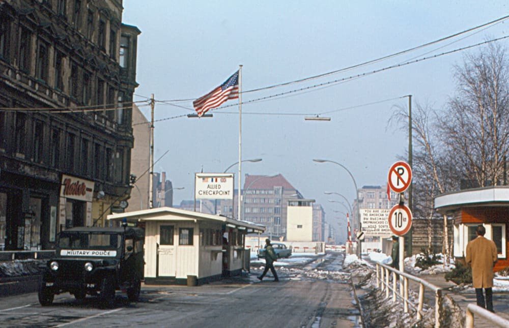 Colour photo of a winter street: a white Allied checkpoint hut under a tall US flag and an 'Allied Checkpoint' sign, a US 'Military Police' jeep on the left, a soldier crossing the road and the East German control area further down the street.