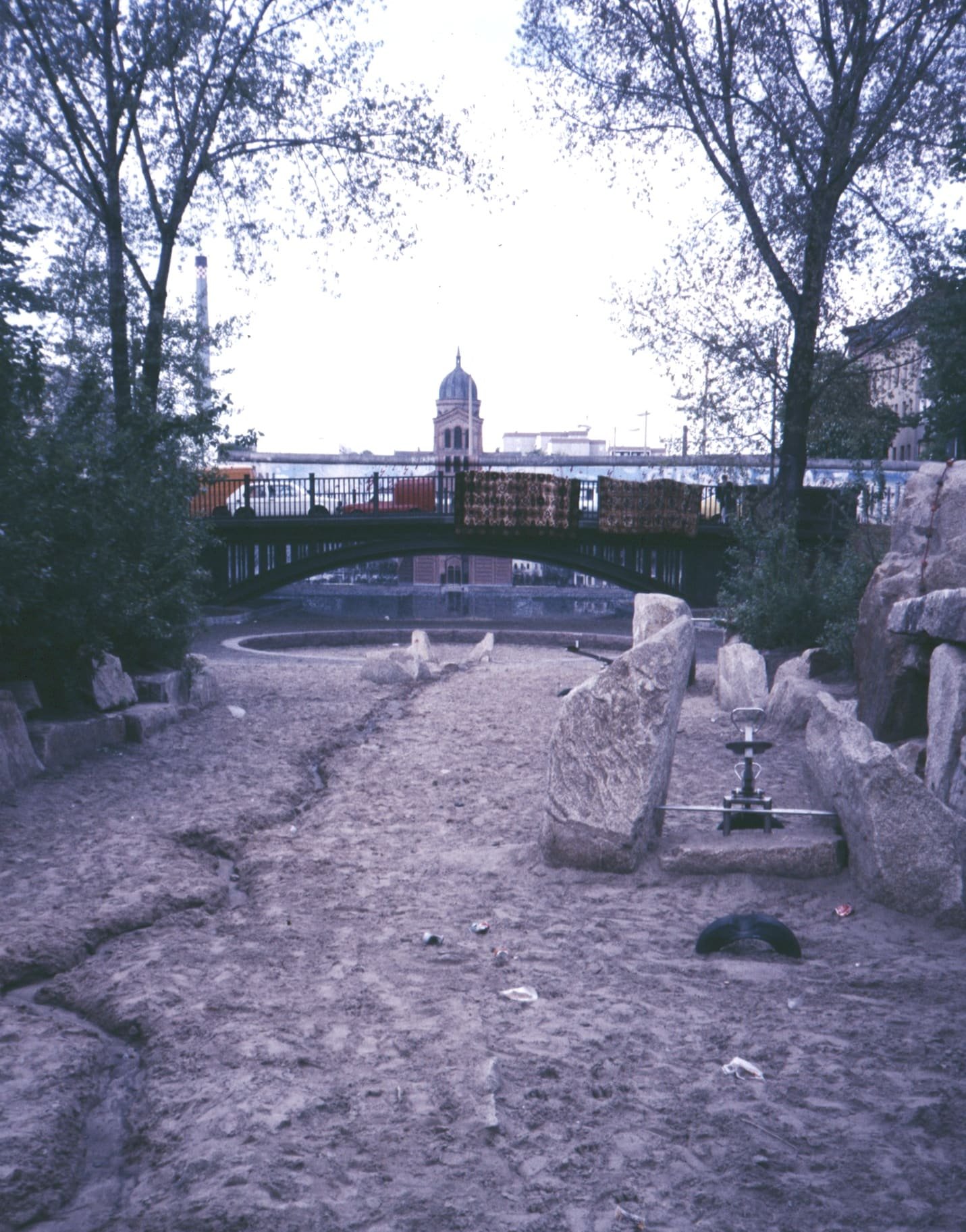Colour photo along the dry, sandy bed of a former canal laid out as a playground with boulders, leading to an arched iron bridge; behind the bridge runs the Wall, with a domed church tower and a tall chimney beyond.