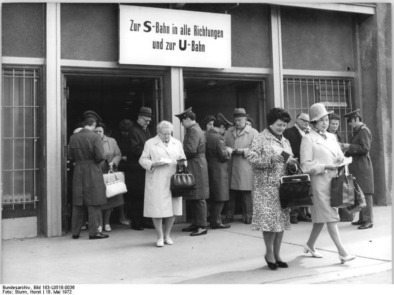 Black-and-white photo: men and women in coats, several holding documents, come out of doorways beneath a sign reading 'Zur S-Bahn in alle Richtungen und zur U-Bahn', with uniformed officials among them.