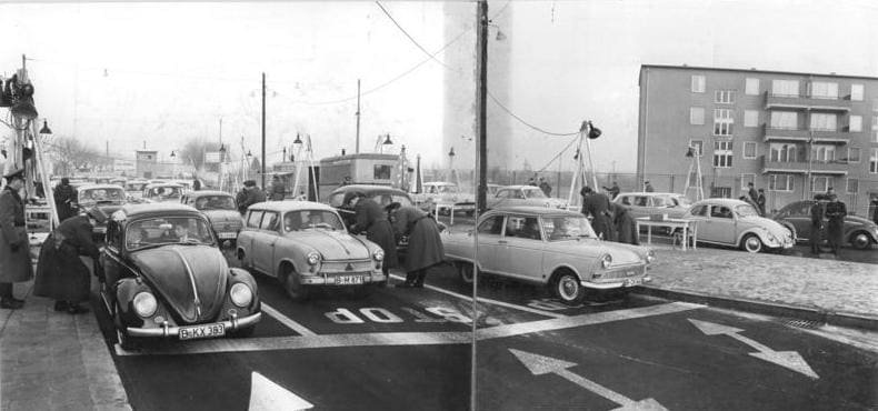 Black-and-white panorama of cars lined up in marked lanes at a checkpoint, with uniformed guards leaning into car windows and a block of flats at right.
