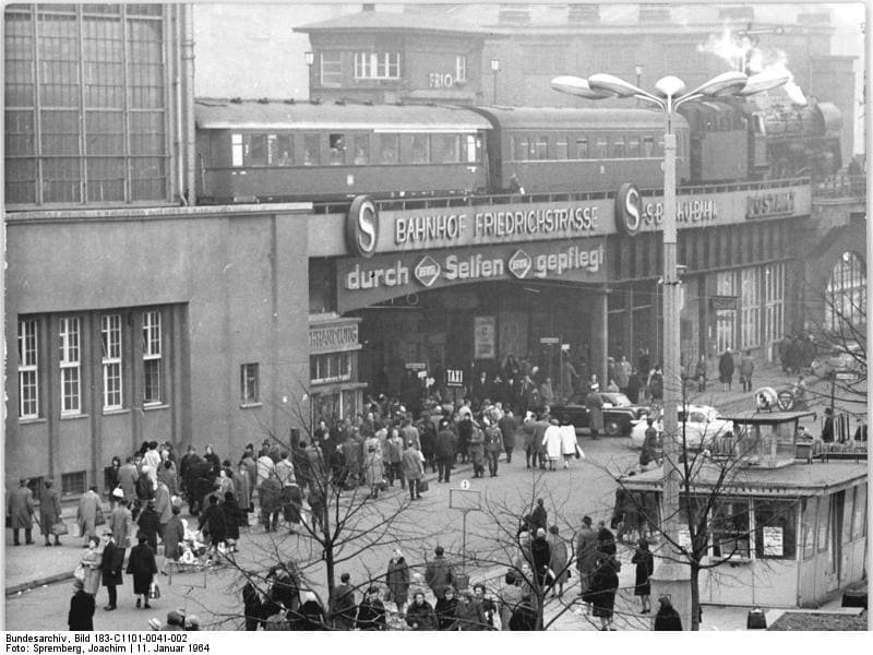 Black-and-white photo of a crowd in front of a station building with an elevated platform above carrying a train, under a large sign reading 'Bahnhof Friedrichstrasse'.