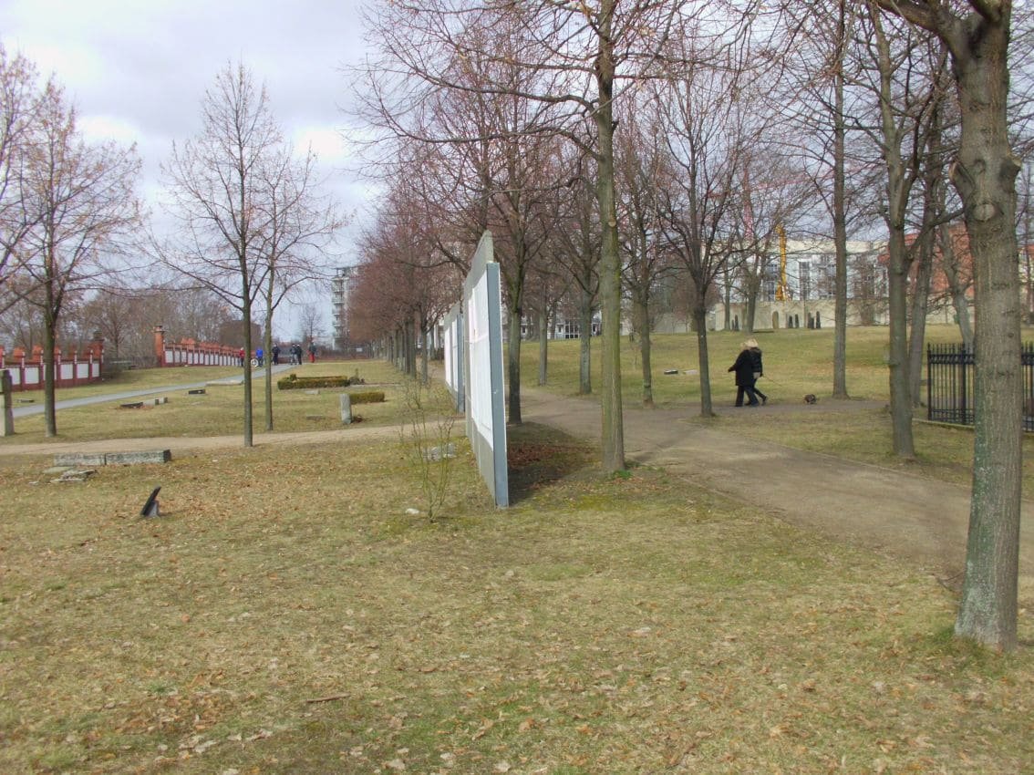 A white concrete wall section standing among bare trees in the grassy cemetery grounds, with a person walking along a path.