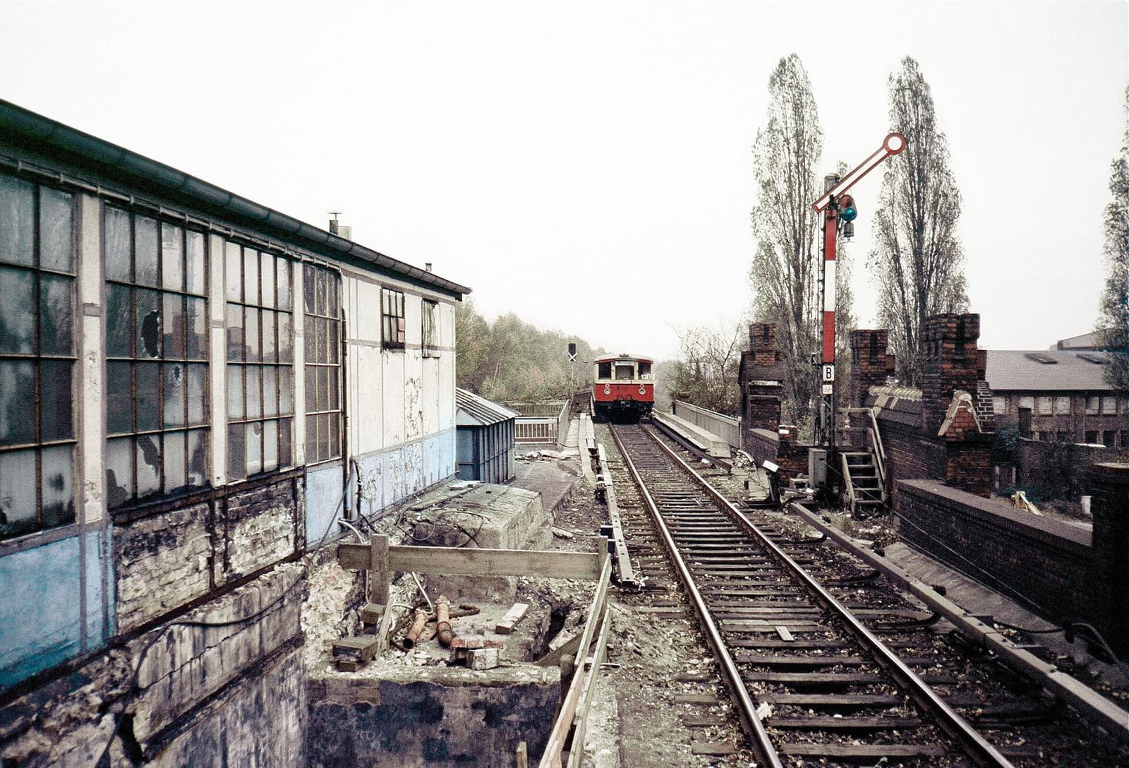 Colour photo along a single railway track on an embankment: a red and cream S-Bahn train approaches past a derelict glazed station building on the left and a semaphore signal and brick walls on the right.