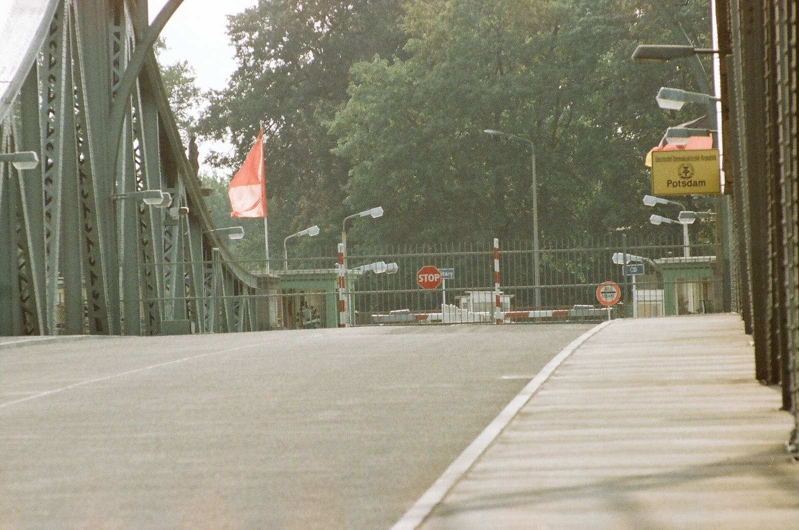 Colour photo along the steel bridge deck towards a barrier gate, a stop sign, a red flag and a yellow 'Potsdam' sign.