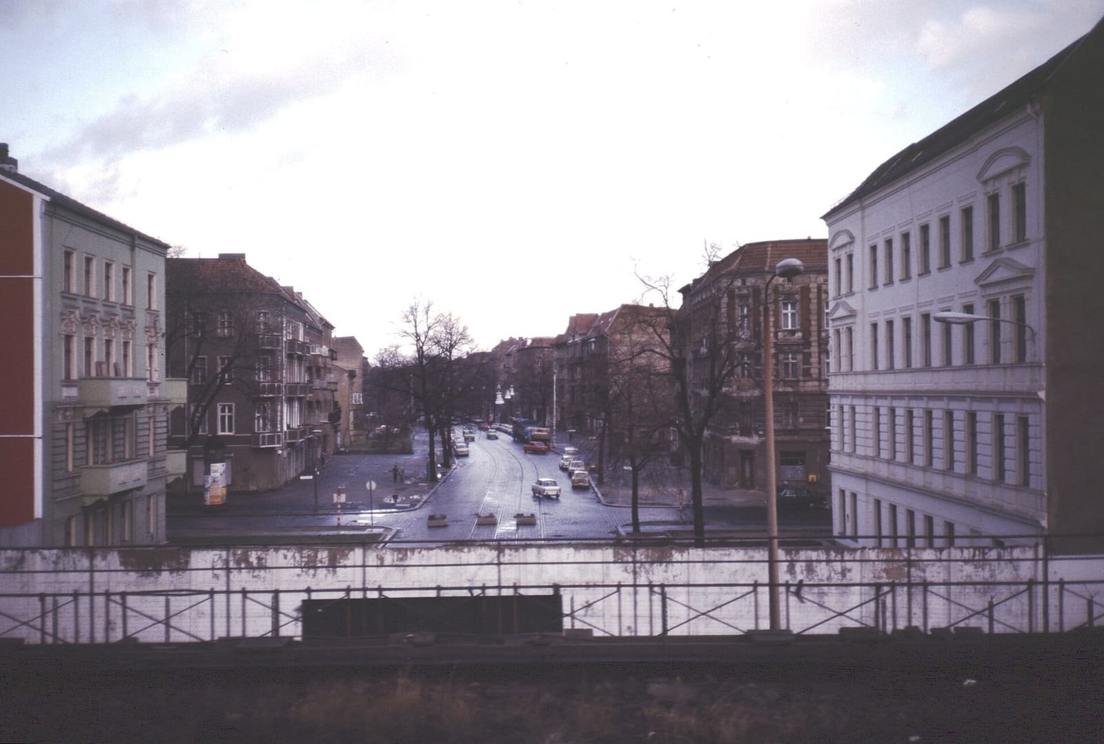 Colour photo taken from a railway bridge over a white wall and a metal fence: a wide street with a tram track runs away between old apartment buildings and bare trees in East Berlin.