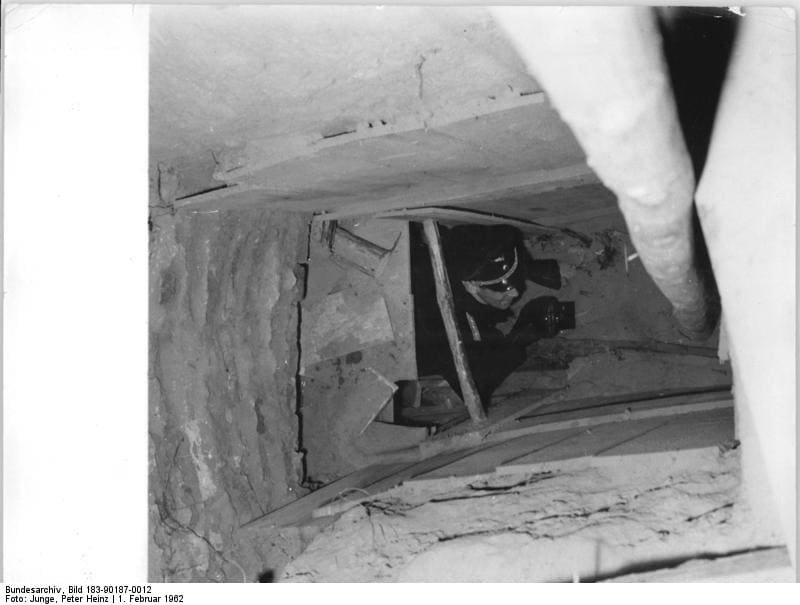 Black-and-white photo looking down a narrow shaft with rough earth walls and timber boards into a dark tunnel.
