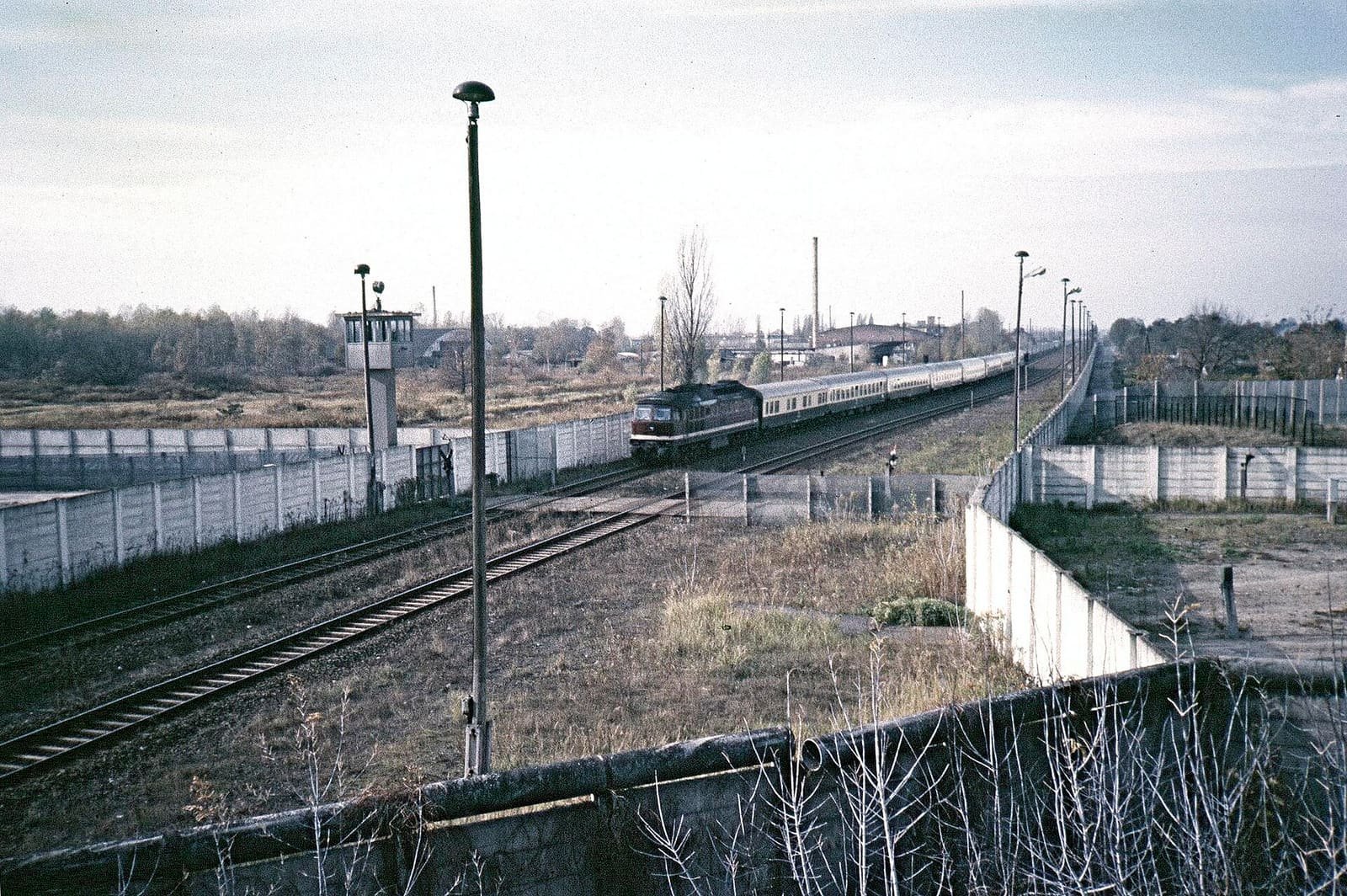Colour photo: a diesel locomotive pulls a long train of passenger coaches along tracks between two lines of white concrete wall; a watchtower stands on the left, tall lamp posts line the tracks and dry grass and weeds fill the foreground.