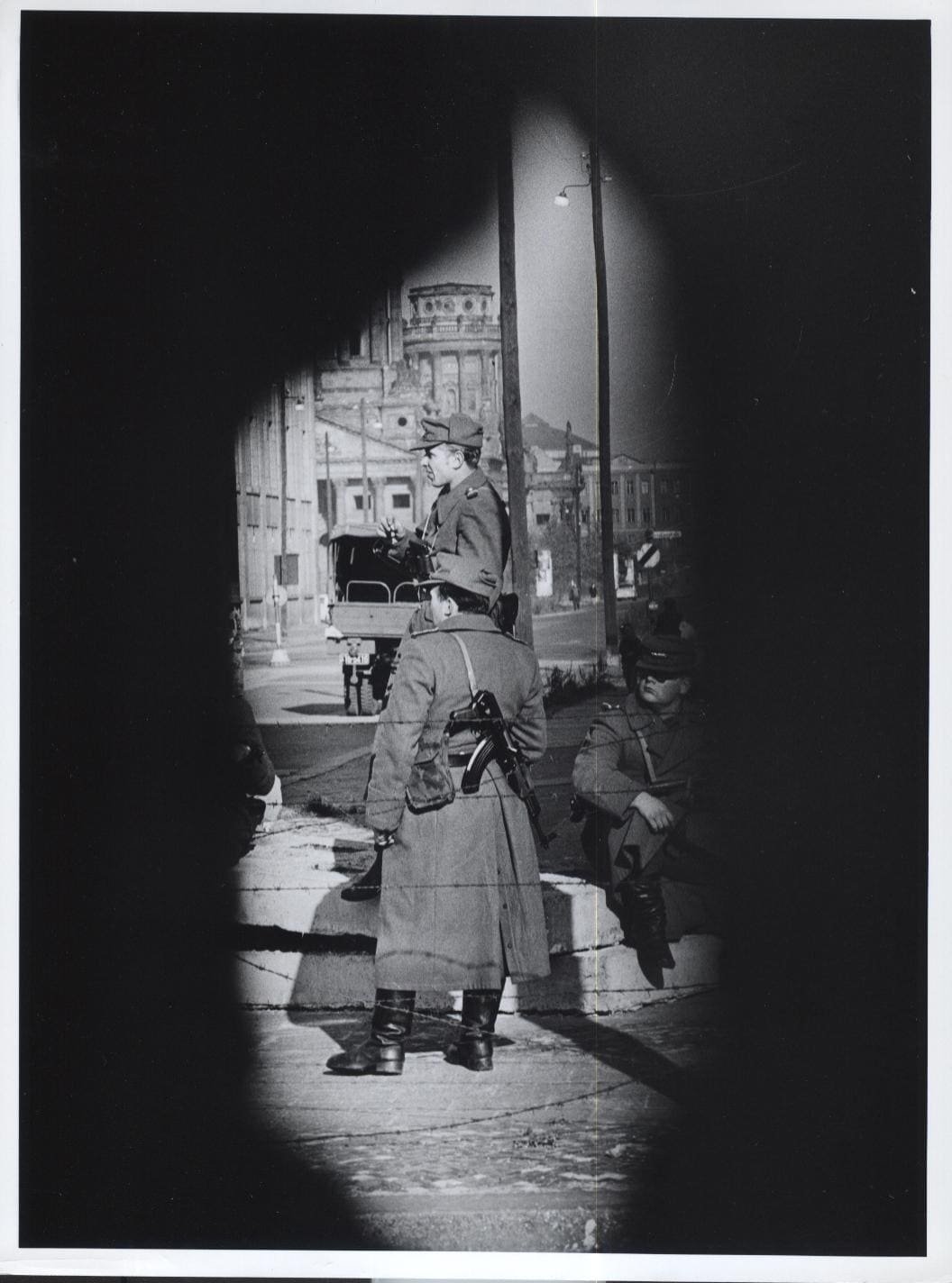 Black-and-white photo framed by the dark edges of a hole in a wall: East German police officers in long coats with submachine guns stand behind barbed wire on a street corner; a lorry and a domed church tower are in the background.