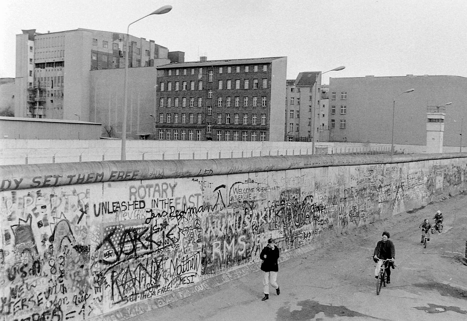 Black-and-white photo of a graffiti-covered wall running along a street, a pedestrian and cyclists on the western side, and behind the wall a second wall, a watchtower and East Berlin buildings.