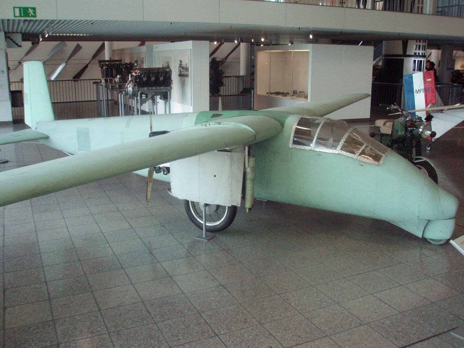 A pale green, homemade single-engine aircraft with long wings, a bubble canopy and a pusher propeller, displayed on a tiled museum floor.