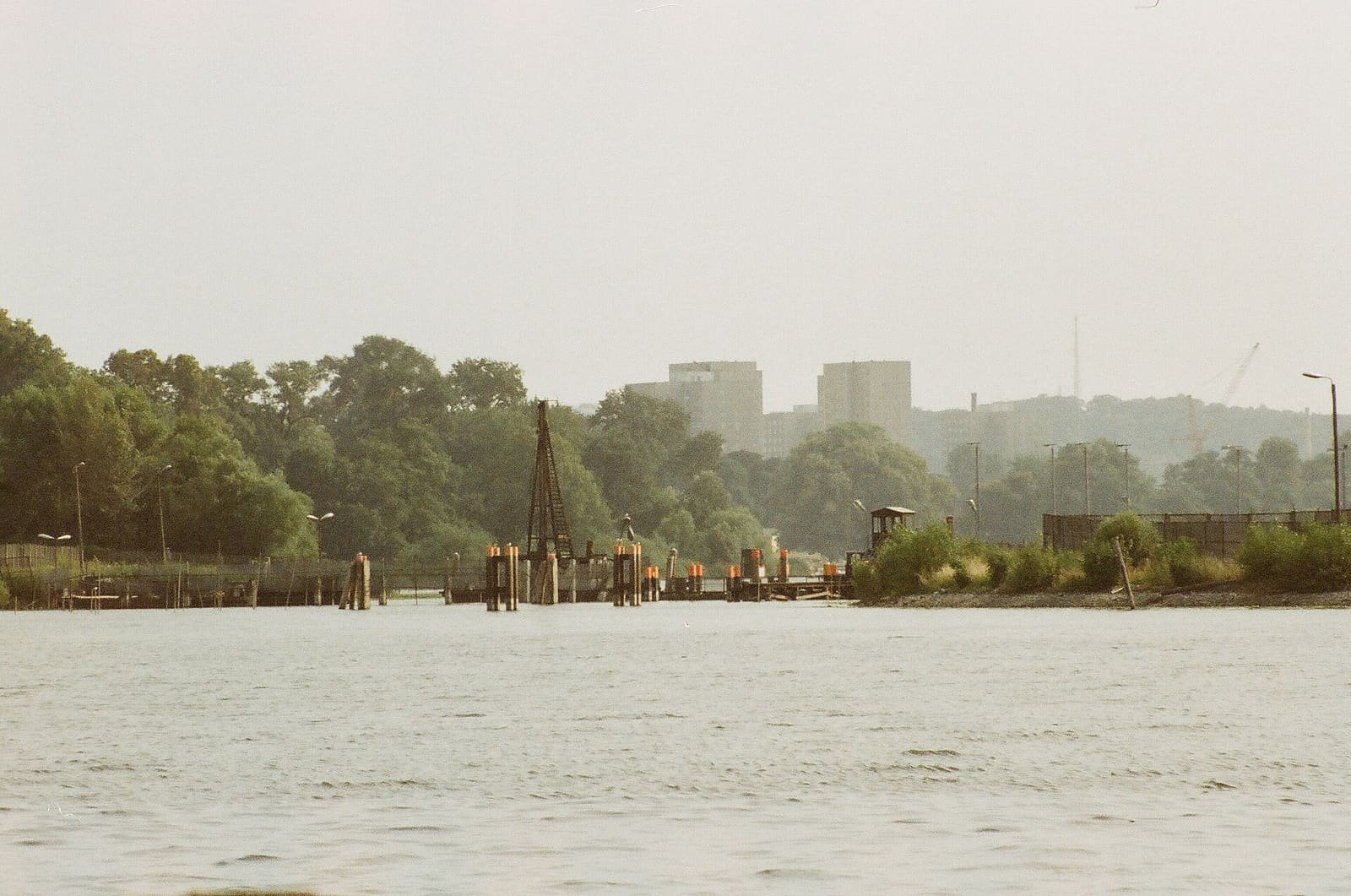 Colour photo taken from the water: a broad grey lake with a line of piles, orange-marked posts and a crane-like structure across it, a small guard hut on the right bank and trees and tower blocks behind.