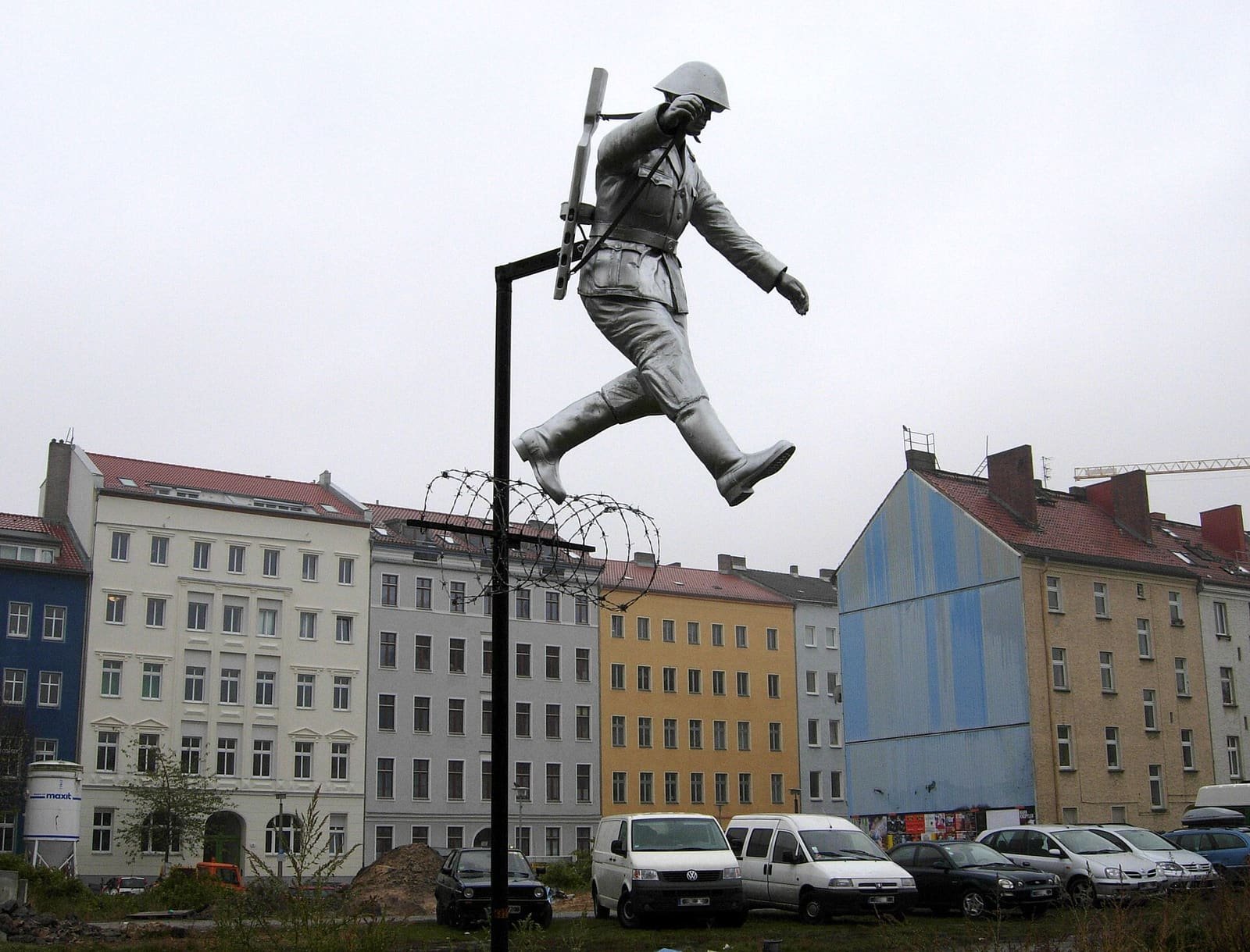 A silver statue of a soldier in helmet and long coat, rifle slung on his back, leaping over a coil of barbed wire on a post, against a grey sky and a row of apartment buildings.