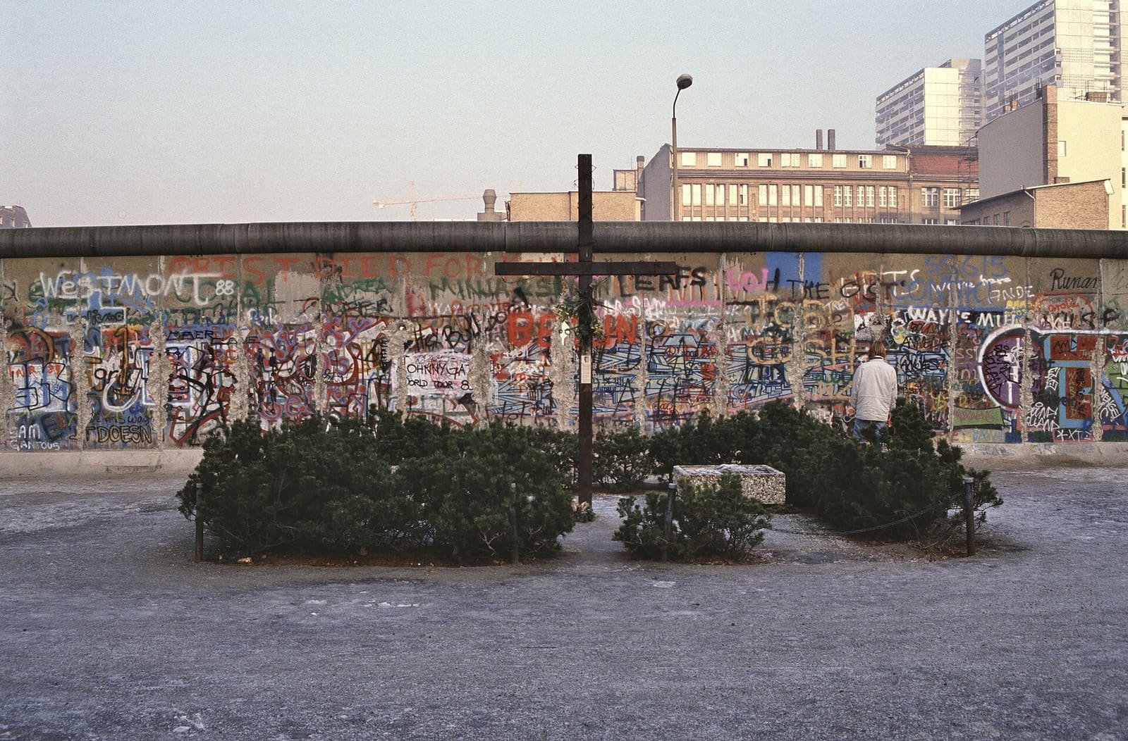 A tall dark wooden cross surrounded by low evergreen shrubs stands in front of a long, colourfully graffitied section of the Berlin Wall, with East Berlin buildings and tower blocks behind.