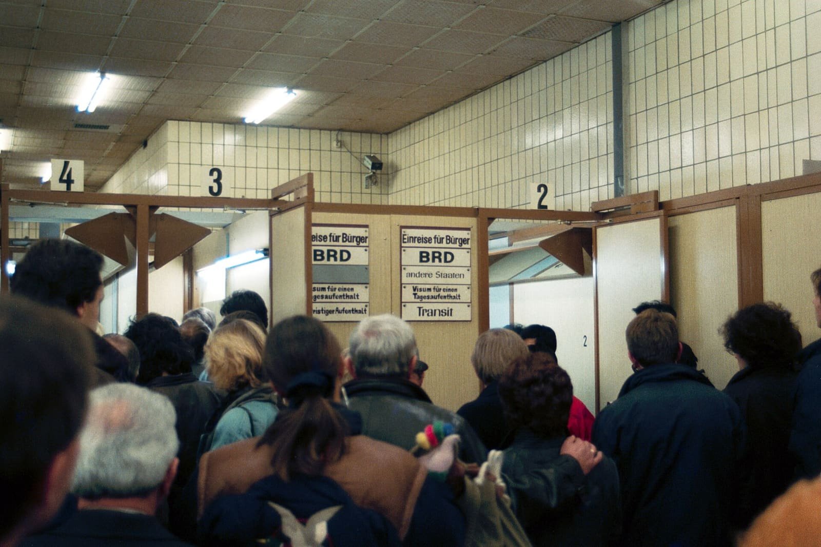 Crowd of people queuing in a tiled hall before numbered wooden control booths with signs reading 'Einreise für Bürger BRD', 'andere Staaten', 'Visum' and 'Transit'.