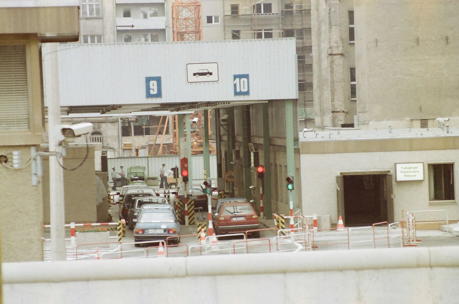 Colour photo of a border checkpoint: cars queue under a metal canopy marked with lane numbers 9 and 10 and a car symbol, beside barriers, traffic lights, a surveillance camera and guards.