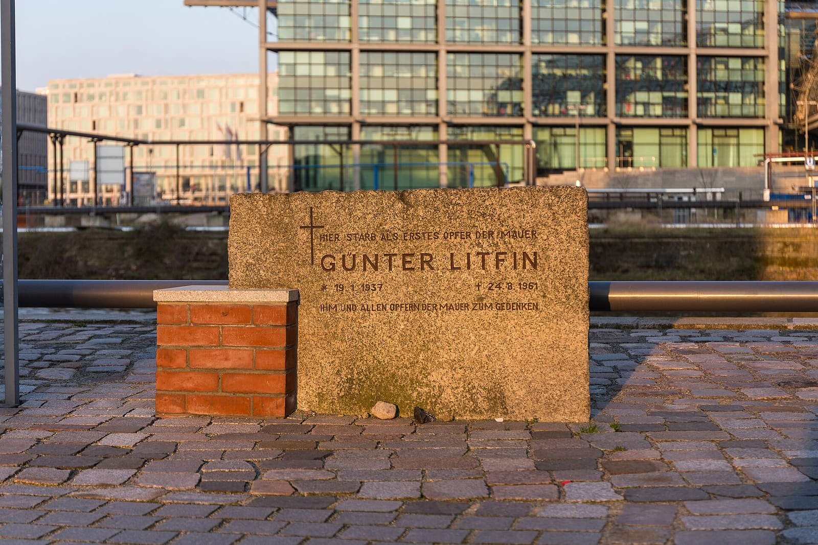 A rough granite block beside a small brick pillar on a cobbled quay, inscribed 'Hier starb als erstes Opfer der Mauer Günter Litfin, 19.1.1937 – 24.8.1961', with a railing, water and modern glass buildings behind.