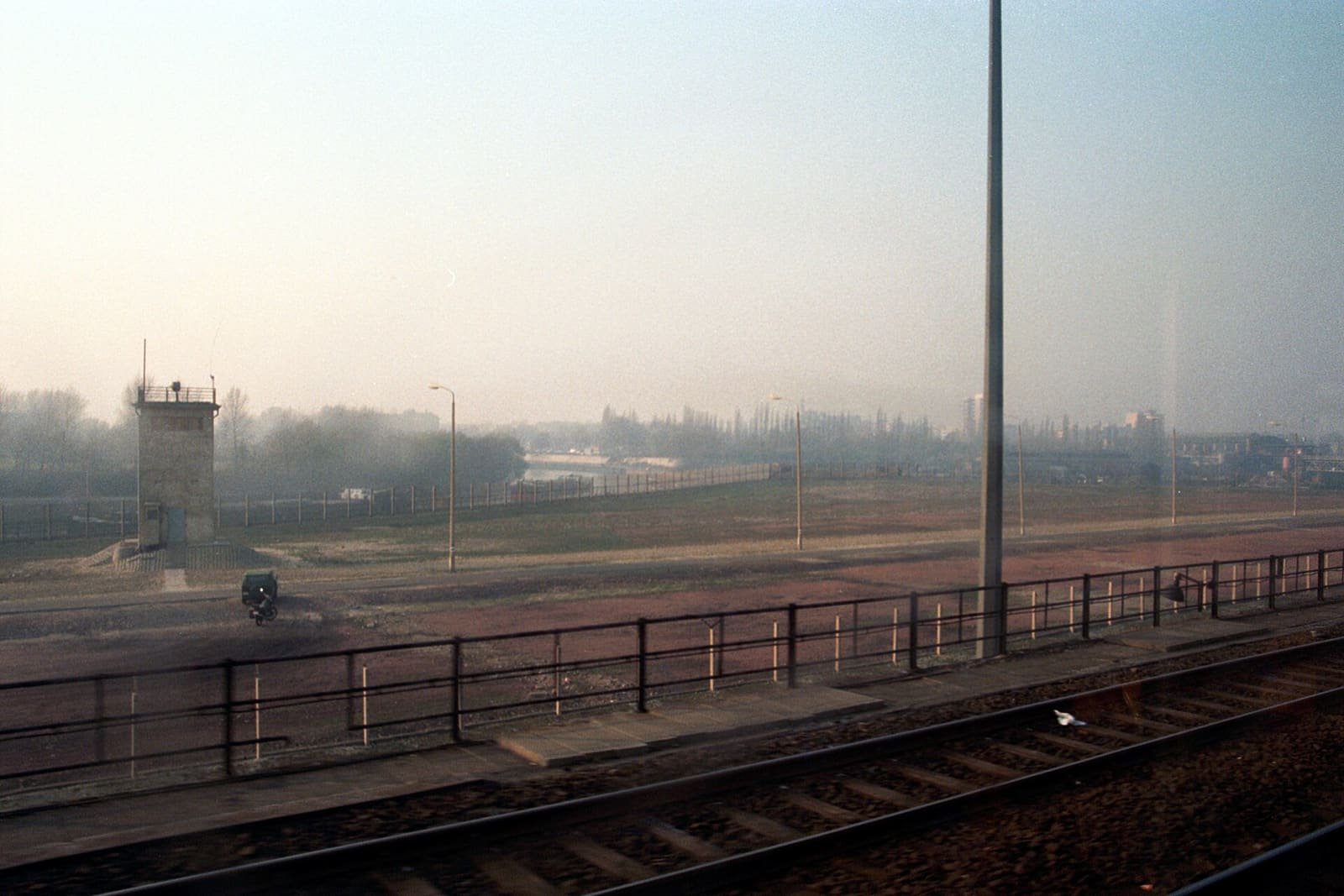 View from a train window across railway tracks and a fence to a wide empty strip of land with a square watchtower, lamp posts and a distant fence in morning haze.